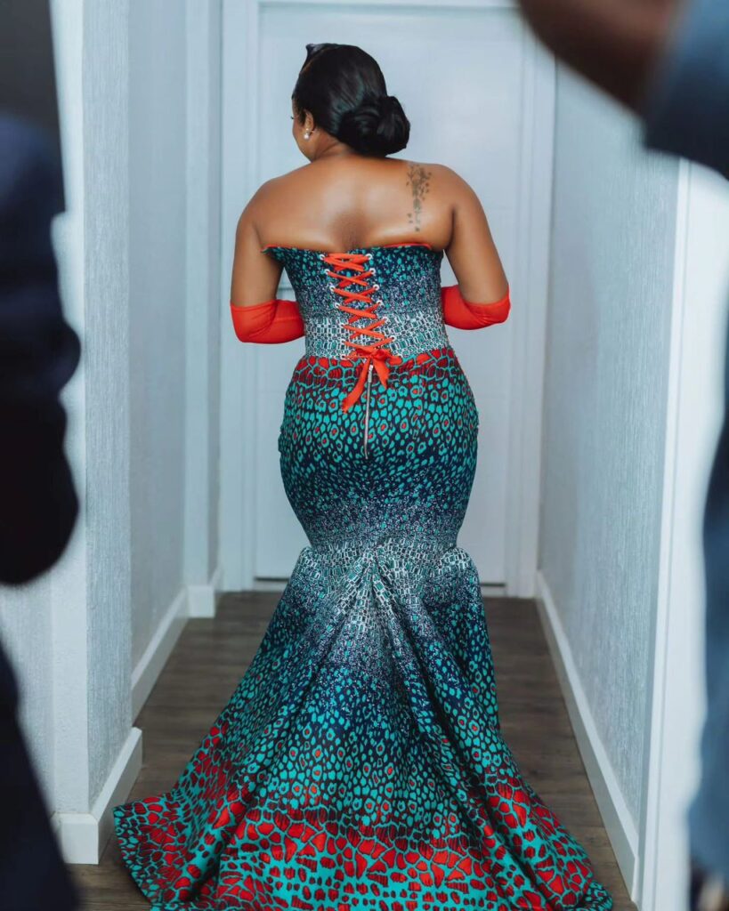 Doreen Avio in a stunning multicolored gown with a long train and a corset-style back, showcasing vibrant patterns as she walks down a hallway.